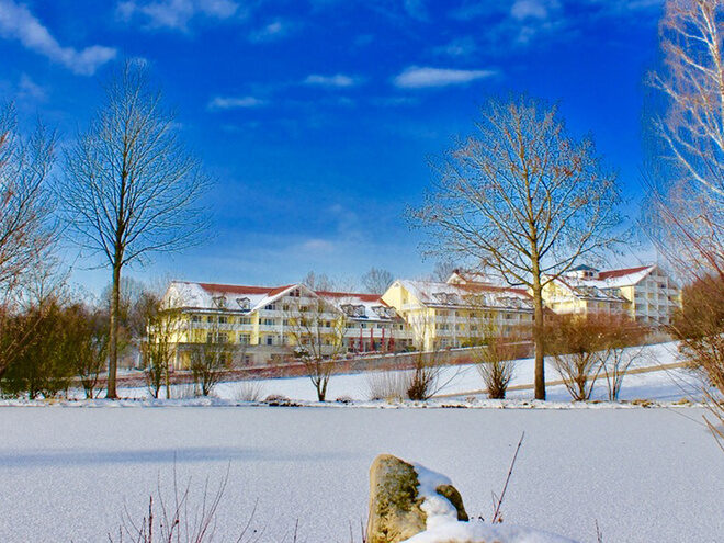 A multi-story building with a snow-covered roof sits behind leafless trees in a wintry landscape, under a bright blue sky, likely representing a clinic and hotel in Bavaria.