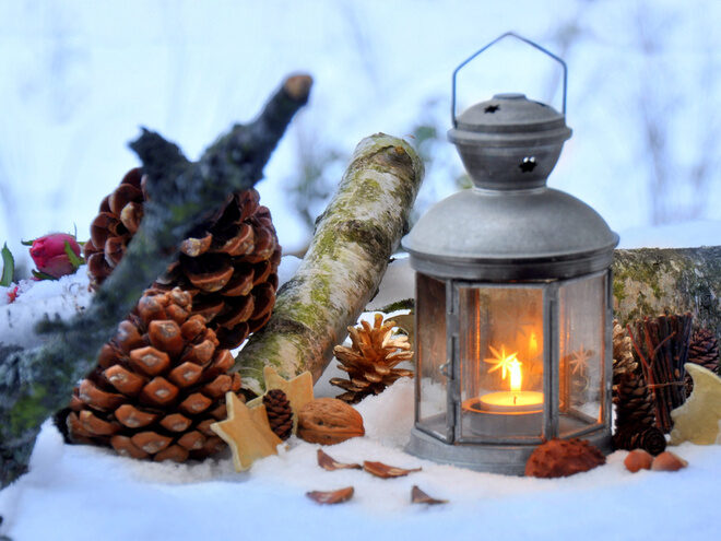 A metal lantern with a glowing candle inside sits on snow, surrounded by pine cones, a birch log, and star-shaped decorations, creating a warm, wintry, outdoor scene.