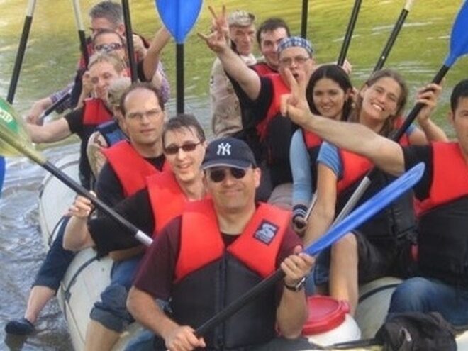 A group of smiling people wearing red life vests hold paddles while sitting in a white raft.